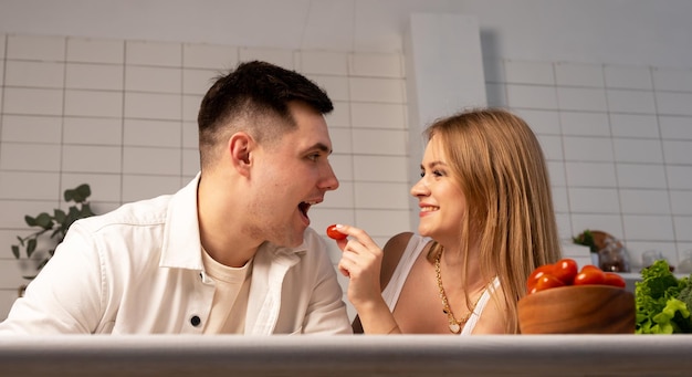 Casal feliz cozinhando em casa linda mulher alimentando homem bonito tomate relacionamento romântico e terno