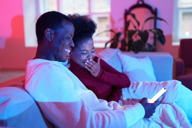 Casal feliz da família africana rindo enquanto relaxa no sofá e olhando para a tela do smartphone