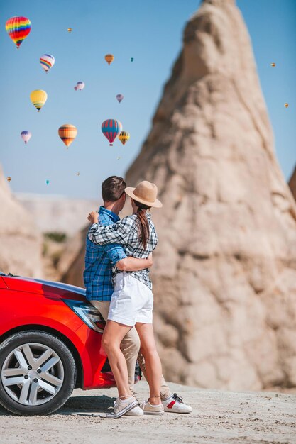 Casal feliz de férias de carro na Capadócia com balões de ar quente ao fundo. Formações de cavernas. Voos de balão de ar quente.