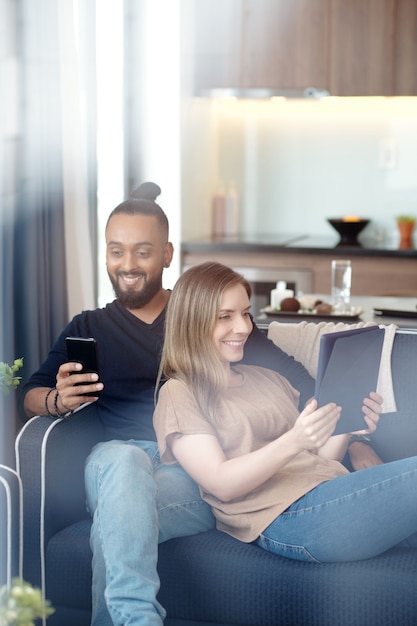 Casal feliz descansando em casa com gadgets em mãos, eles estão assistindo vídeos ou lendo mensagens