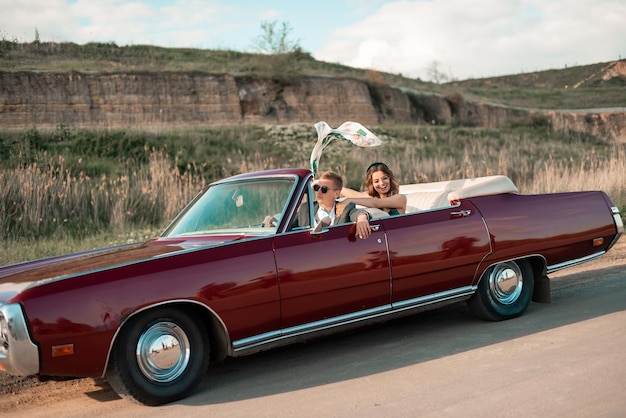 Foto casal feliz dirigindo pela estrada rural ao pôr do sol em carro conversível vintage