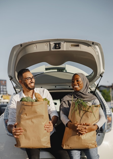 Casal feliz diversificado se divertindo depois de fazer compras. sentado no porta-malas do carro cheio de alimentos frescos e saudáveis.