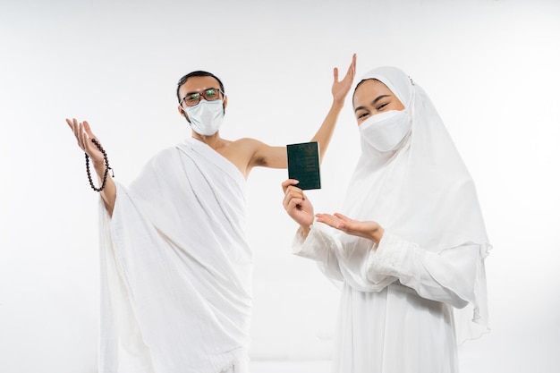 Casal feliz em roupas de ihram e máscara segurando um passaporte em pé em um fundo isolado