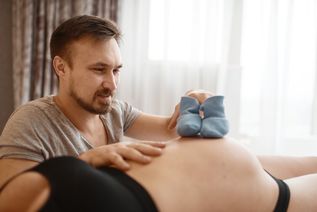 Casal feliz, esposa grávida e marido brinca com roupas para recém-nascidos na barriga em casa. Gravidez, período pré-natal. Mamãe e papai expectantes estão descansando na cama