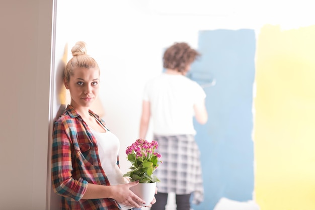 Foto casal feliz fazendo reformas em casa, o homem está pintando o quarto e a mulher segura o pote com flores