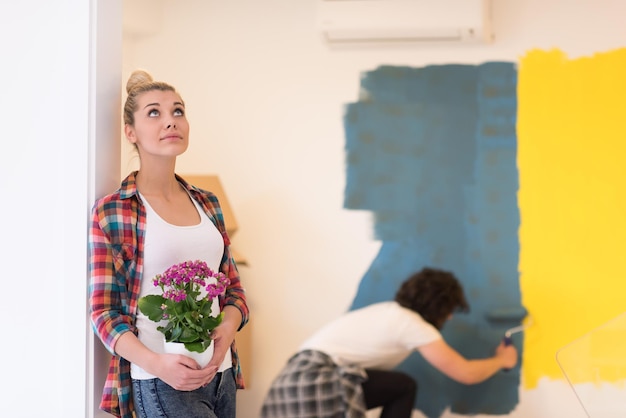 Foto casal feliz fazendo reformas em casa, o homem está pintando o quarto e a mulher segura o pote com flores