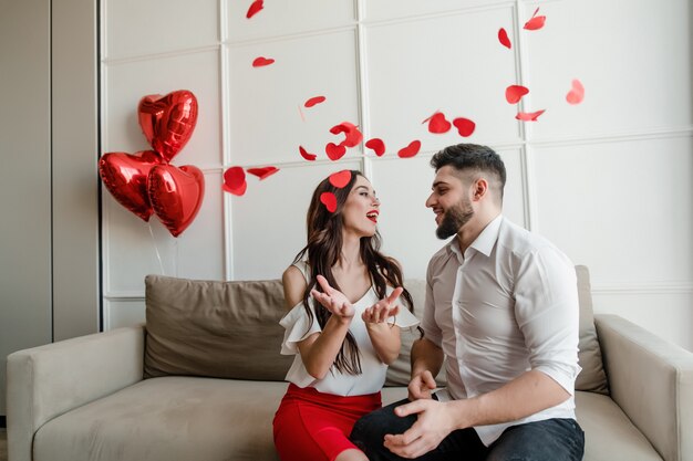 Foto casal feliz homem e mulher apaixonada por confetes em forma de coração no ar, sentado no sofá em casa