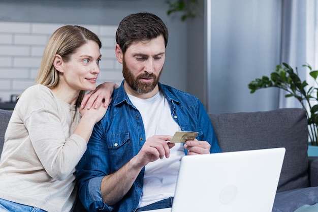 Casal feliz homem e mulher sentados em casa no sofá usando laptop para compras on-line segurando um cartão de banco de crédito