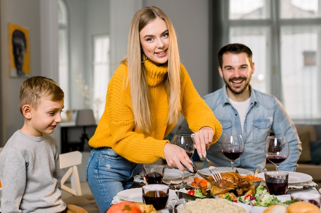 Foto casal feliz pais e filho pequeno sentado na mesa festiva e vai comer peru assado. família feliz, jantando delicioso juntos em casa, enquanto a mãe corta a turquia