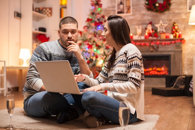 Casal feliz pensando nos presentes que eles têm que comprar para o Natal usando seu laptop em frente à lareira. Taça de champanhe.