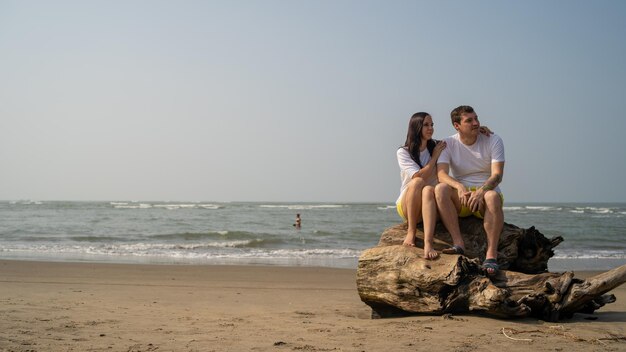 Casal feliz posando em troncos perto do mar Casal apaixonado abraçando durante o encontro na praia contra o mar acenando e céu sem nuvens