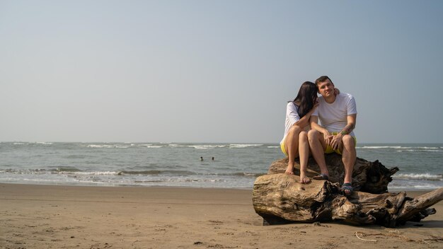 Casal feliz posando em troncos perto do mar Casal apaixonado abraçando durante o encontro na praia contra o mar acenando e céu sem nuvens