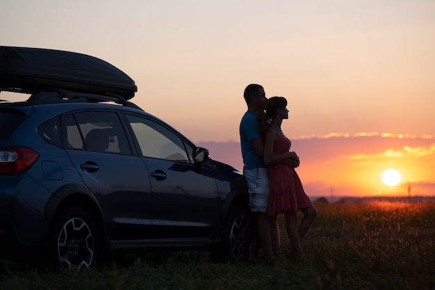 Casal feliz relaxando ao lado de seu carro SUV durante a viagem de lua de mel ao pôr do sol. Jovem e mulher, aproveitando o tempo juntos viajando de veículo.