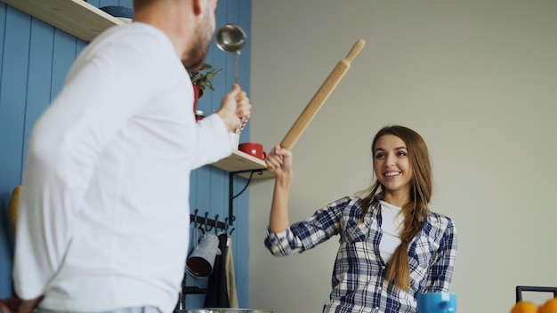 Casal feliz se divertindo na cozinha esgrima com concha e rolo enquanto cozinha o café da manhã