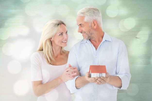 Casal feliz segurando casa modelo em miniatura contra círculos de luz no fundo cinza