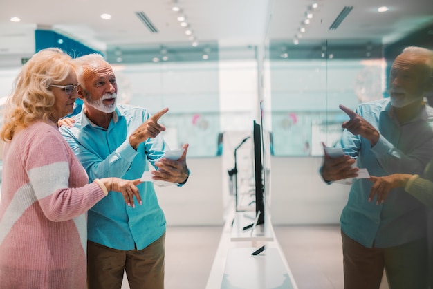 Casal feliz sênior, escolhendo qual aparelho de televisão para comprar. Homem apontando para tv de plasma. Interior da loja de tecnologia.