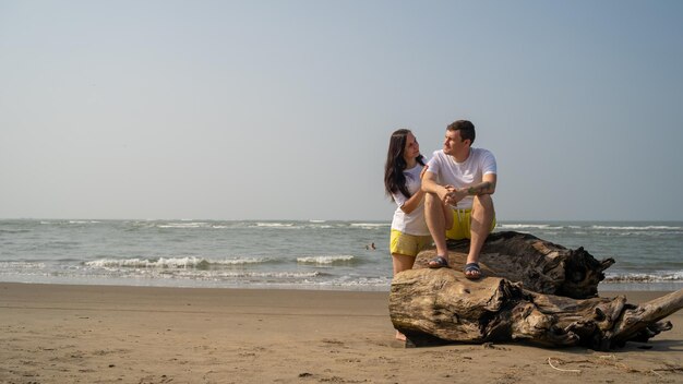 Casal feliz sentado em troncos perto do mar Casal apaixonado abraçando e olhando um ao outro enquanto está sentado no tronco durante o encontro na praia contra o mar acenando e céu sem nuvens