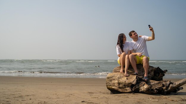 Casal feliz sentado em troncos perto do mar Casal apaixonado abraçando e tirando selfies enquanto está sentado no tronco durante o encontro na praia contra o mar acenando e céu sem nuvens