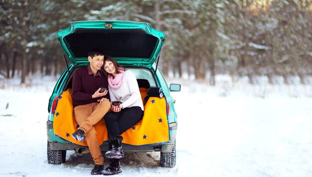 Casal feliz sentado no porta-malas aberto de um carro no inverno ao ar livre com canecas de chá quente nas mãos