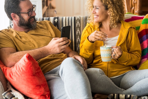 Casal feliz sentado no sofá e olhando um para o outro enquanto bebe café e usa o telefone celular em casa. Casal apaixonado, passando momentos de lazer juntos em casa. Casal olhando cara a cara.