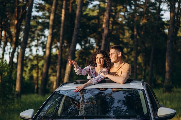 Casal feliz sentado no topo do telhado da minivan ao pôr do sol Jovens se divertindo nas férias de primavera viajando ao redor do mundo Travellove e conceito de férias Foco nos rostos