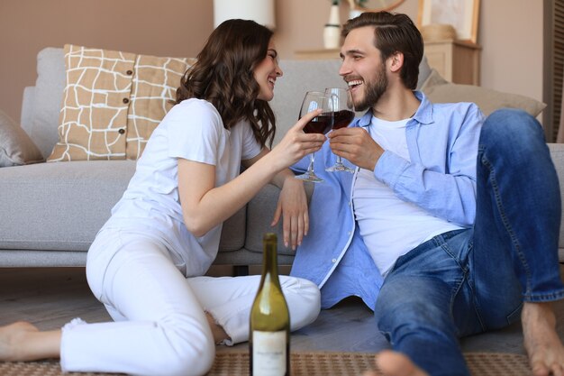 Casal feliz sentado, relaxando no chão da sala de estar, bebendo vinho tinto. Sorridente e jovem marido e mulher descansam em casa para desfrutar de um encontro romântico no fim de semana familiar juntos.