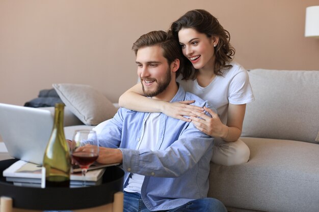 Casal feliz sentado, relaxando no chão da sala de estar, usando o laptop para fazer compras online juntos, assistindo filme.