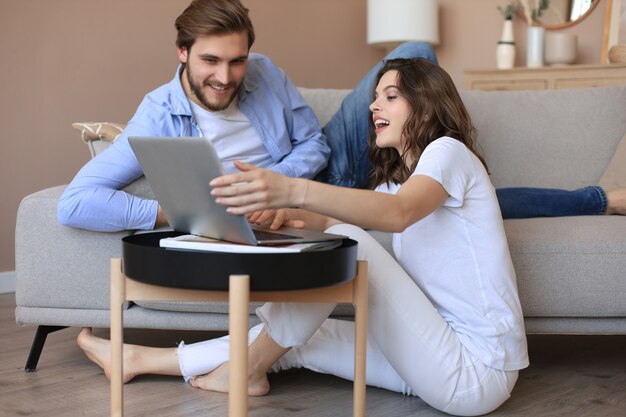 Casal feliz sentado, relaxando no chão da sala de estar, usando o laptop para fazer compras online juntos, assistindo filme.