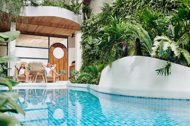 Casal feliz sonhando acordado em espreguiçadeiras à beira da piscina do hotel spa e bebendo coquetéis de frutas