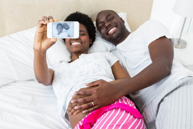 Casal feliz tocando a barriga de grávida tomando selfie na cama