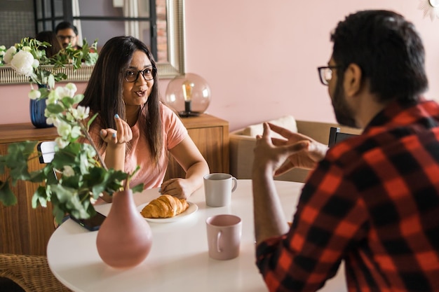 Foto casal feliz tomando café da manhã e conversando na mesa de jantar de manhã menina indiana e cara latino conceito de relacionamento e diversidade