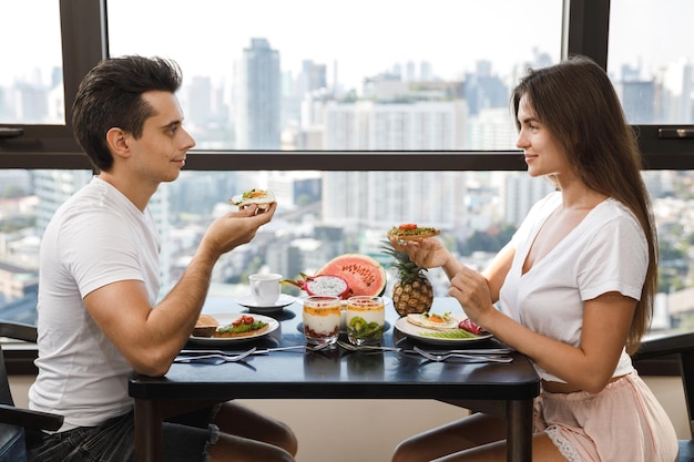 Casal feliz tomando café da manhã saudável em apartamento moderno com grandes janelas e vista da cidade