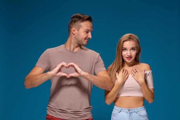 Casal feliz vestiu roupas casuais, forma de coração com os dedos, retrato de estúdio sobre fundo azul