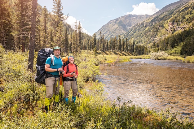 Casal feliz viajantes com bastões de trekking e uma mochila grande caminha ao longo de uma trilha em um parque nacional com um rio de montanha ao fundo