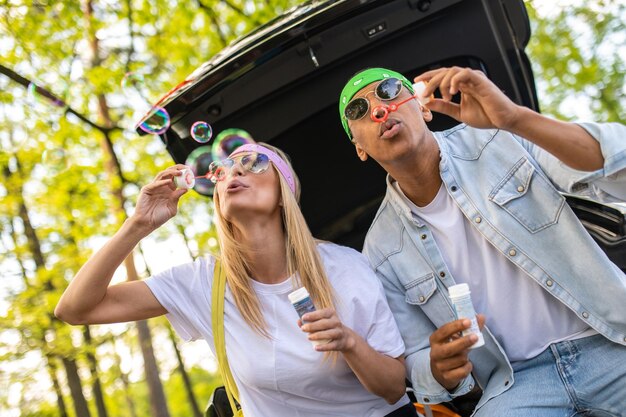 Foto casal fofo sentado no porta-malas do carro e soprando bolhas