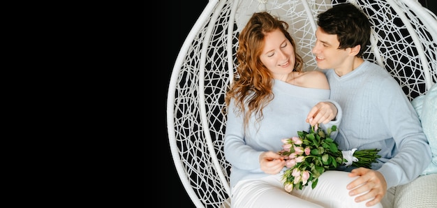 Casal garota e cara abraçando em uma cadeira grande com um buquê de rosas. Suéter branco e azul.