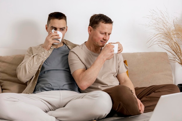 Casal gay feliz com roupas casuais passando tempo juntos em casa assistindo filme no laptop e bebendo chá Dois homens caucasianos relaxando relacionamentos homossexuais e amor alternativo