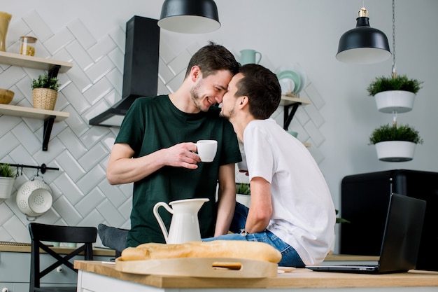 Casal gay feliz tomando café na cozinha e usando o laptop. Gays amorosos se beijando, cara a cara enquanto passam o lazer em casa