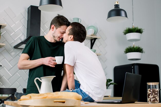Casal gay feliz tomando café na cozinha e usando o laptop. Gays amorosos se beijando, cara a cara enquanto passam o lazer em casa
