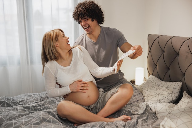 Casal grávida feliz se divertindo enquanto procurava roupas novas para o próximo bebê em casa no quarto.