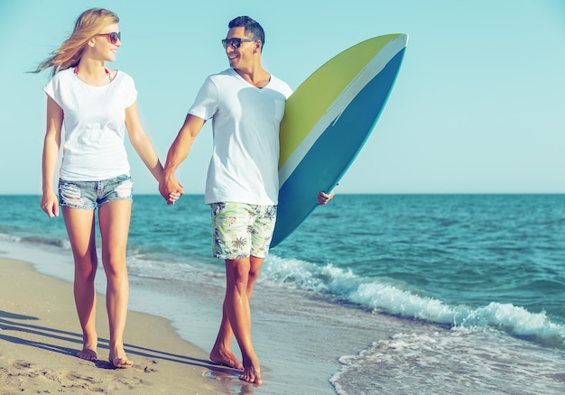 Casal hispânico caminha na praia com prancha de surf se divertindo ao ar livre