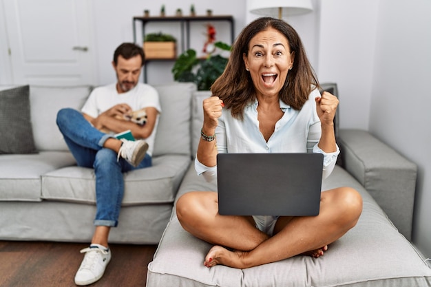 Casal hispânico de meia-idade em casa, mulher usando laptop comemorando surpresa e surpresa pelo sucesso com os braços levantados e olhos abertos. conceito de vencedor.