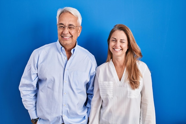 Foto casal hispânico de meia-idade em pé sobre fundo azul piscando olhando para a câmera com expressão sexy rosto alegre e feliz