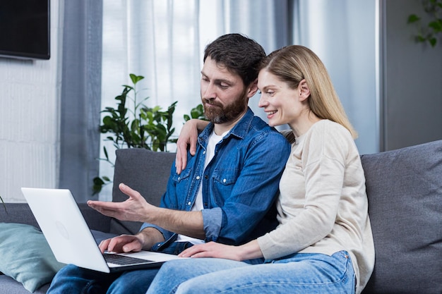 Casal homem e mulher sentados juntos em casa no sofá usando laptop para bate-papo por vídeo e conversando com amigos