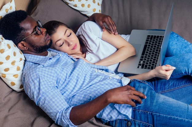 Casal internacional sorrindo olhando para laptop enquanto descansava na cama no quarto juntos em casa