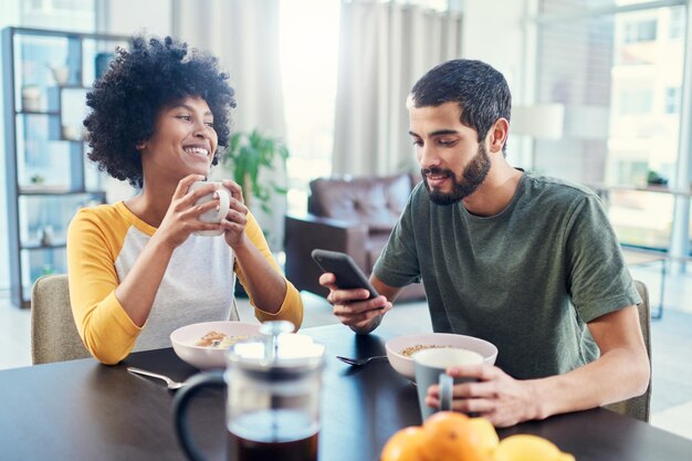 Casal interracial feliz e pequeno-almoço com telefone para a manhã mídia social ou agenda em casa Jovem homem e mulher com sorriso no smartphone móvel para lanche saudável ou dicas de dieta em casa
