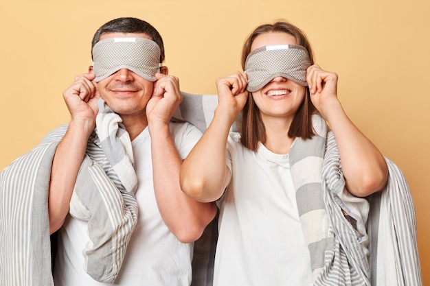 Casal irreconhecível homem e mulher dormindo máscara de olho embrulhado em cobertor isolado sobre fundo bege escondendo os olhos com venda nos olhos sorrindo felizmente