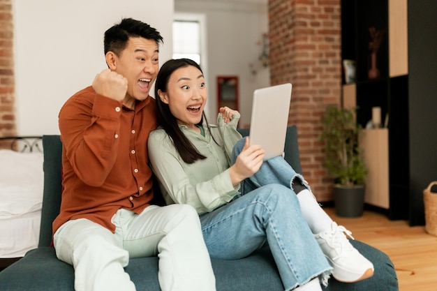 Casal japonês muito feliz sentado no sofá com tablet digital comemorando vitória online no espaço de cópia em casa