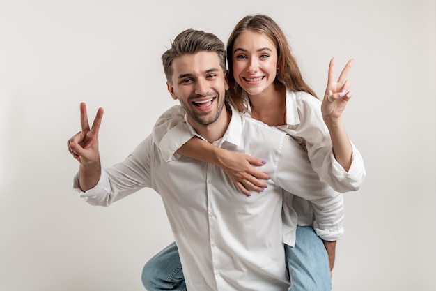 Casal jovem alegre animado em pé isolado sobre um fundo branco, cavalgando nas costas, gesto de paz