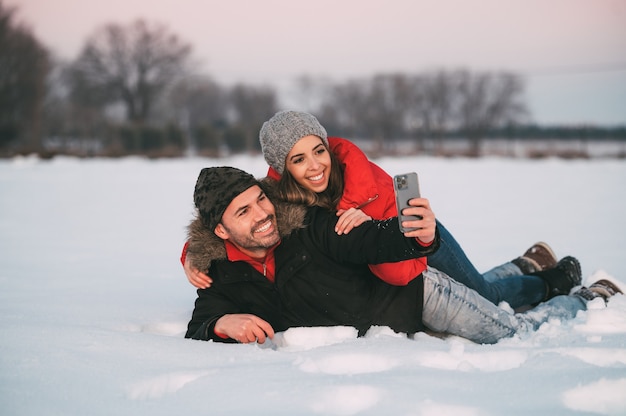 Casal jovem alegre em roupas quentes, deitado na neve e tirando uma selfie no smartphone enquanto se diverte no campo de inverno